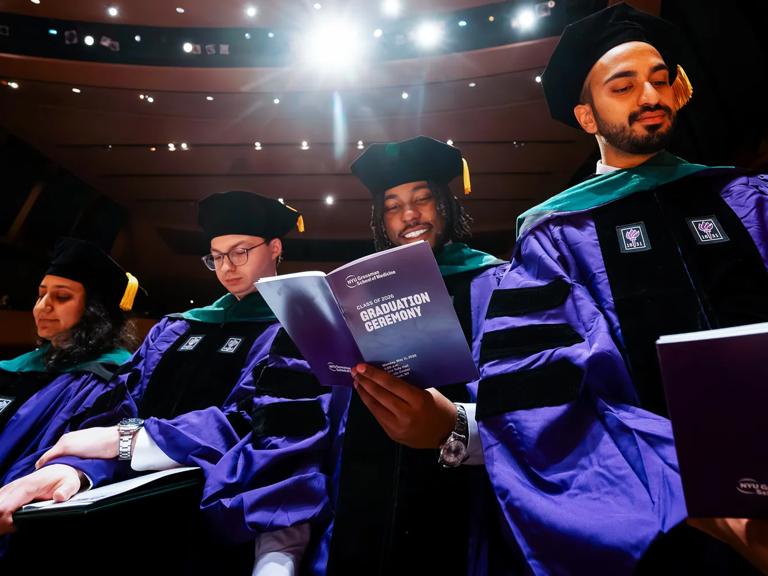 Four Grossman School of Medicine graduates stand during commencement, smiling while they look down at purple graduation ceremony run-of-show pamphlets.