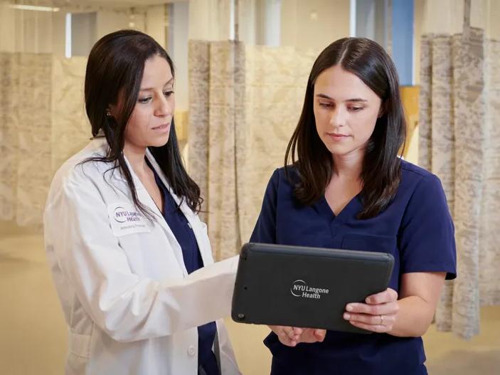A female wearing a white coats talks to another female wearing dark blue hospital scrubs while holding a digital tablet in an NYU Langone hospital.