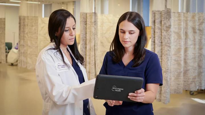 A female wearing a white coats talks to another female wearing dark blue hospital scrubs while holding a digital tablet in an NYU Langone hospital.