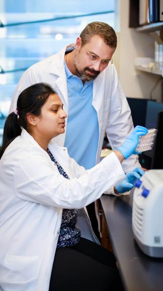 Two people work in a Grossman School of Medicine lab, a female is sitting down and inserts samples into a  machine, while a male colleague stand over her watching.