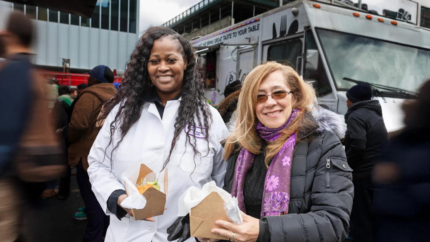 A female doctor and a woman stand next to each other holding containers of food in front of a food truck outside of NYU Langone Hospital—Brooklyn.