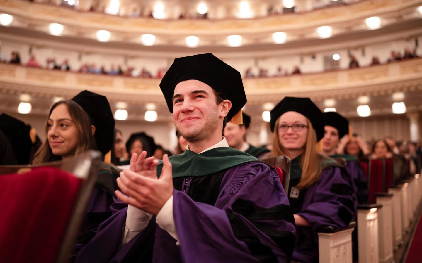 A male Grossman School of Medicine medical student smiles and claps while wearing full graduation regalia with his peers during the annual doctoral hooding ceremony.