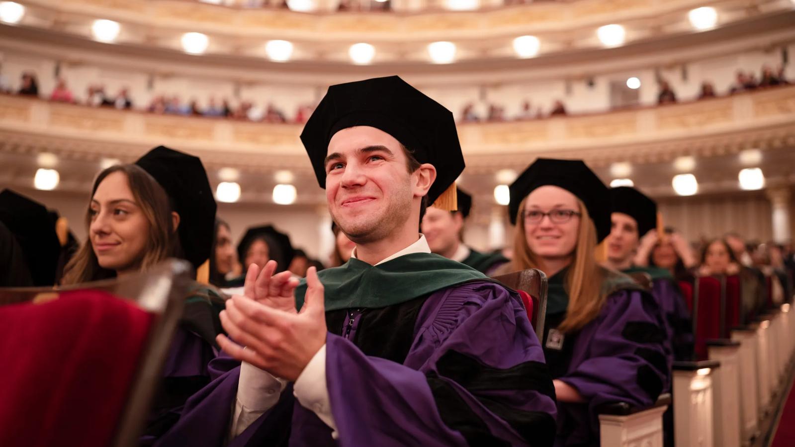 A male Grossman School of Medicine medical student smiles and claps while wearing full graduation regalia with his peers during the annual doctoral hooding ceremony.