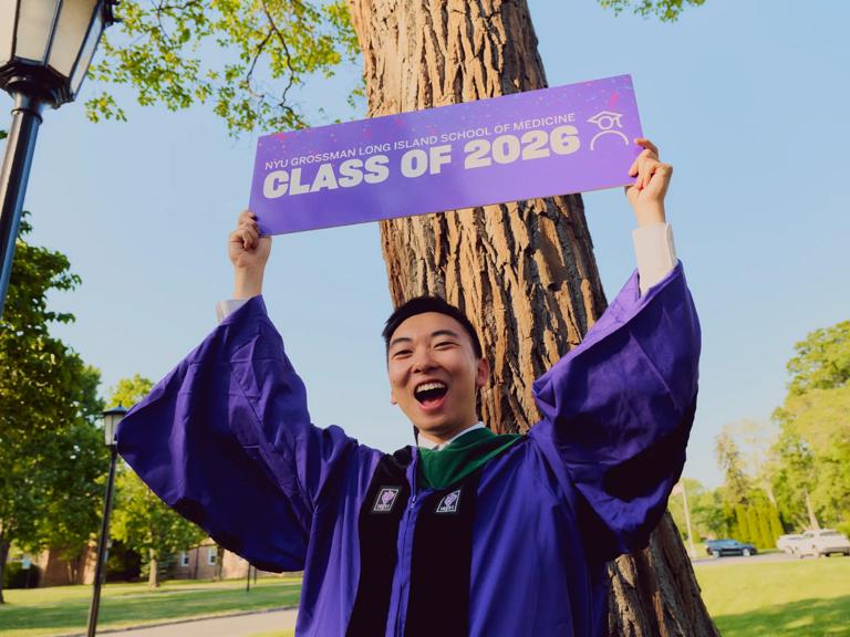 A male NYU Grossman Long Island School of Medicine graduate wearing a purple commencement robe, holds up a purple Class of 2026 banner above his head after his graduation ceremony.