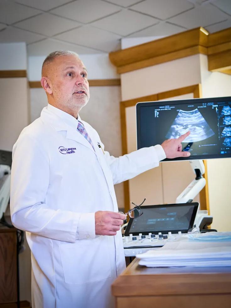 A male NYU Grossman Long Island School of Medicine professor of obstetrics and gynecology explains an ultrasound while pointing at the machine's screen.