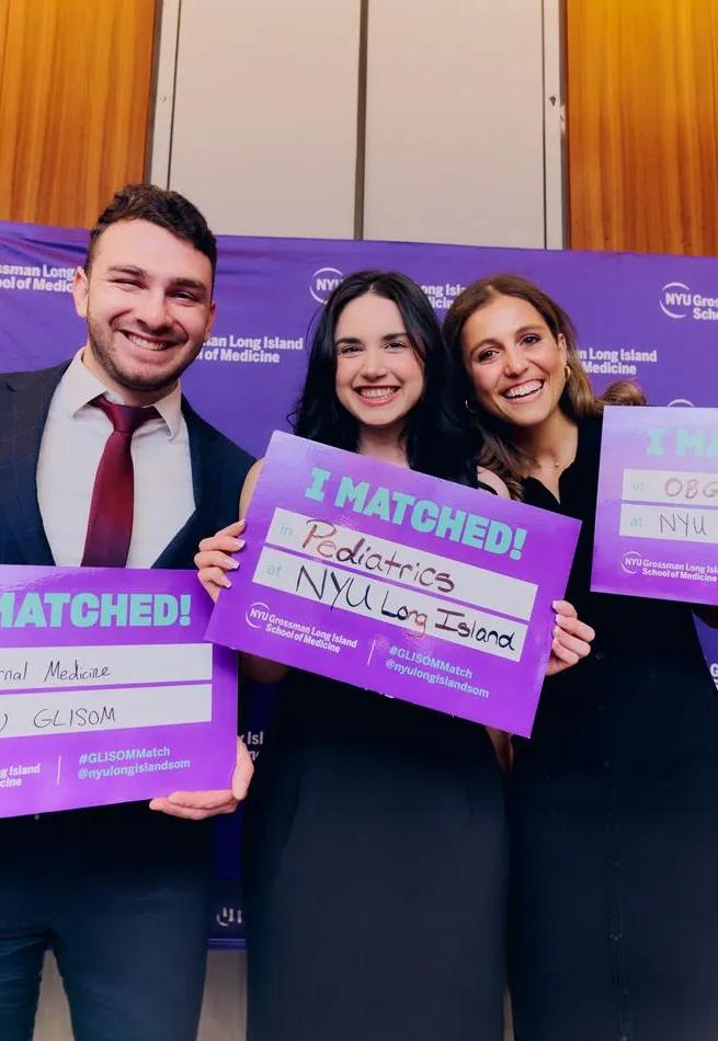 Three people in formal attire stand in front of a purple NYU Grossman Long Island School of Medicine backdrop, holding bright purple “I MATCHED!” signs. The person in the center holds a sign that reads “I MATCHED! in Pediatrics at NYU Long Island,” celebrating residency Match Day. 