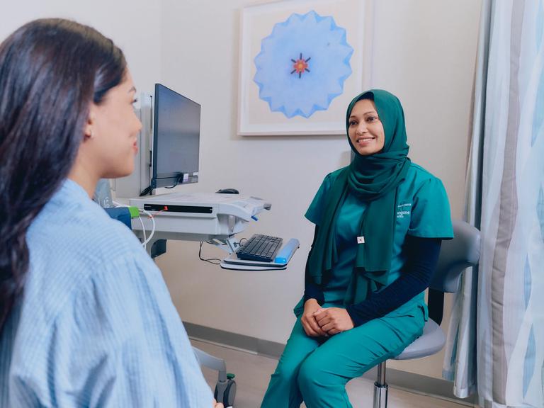 A female medical professional, right, sits in an exam room and smiles at a female patient, left, seated.