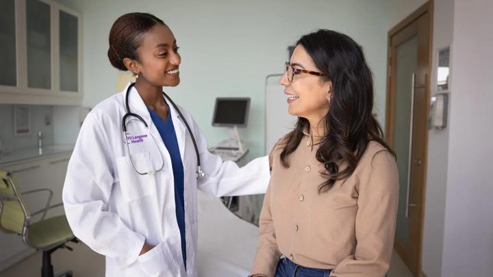 A female doctor, left, stands and puts her hand on a female patient, right, while speaking to her.