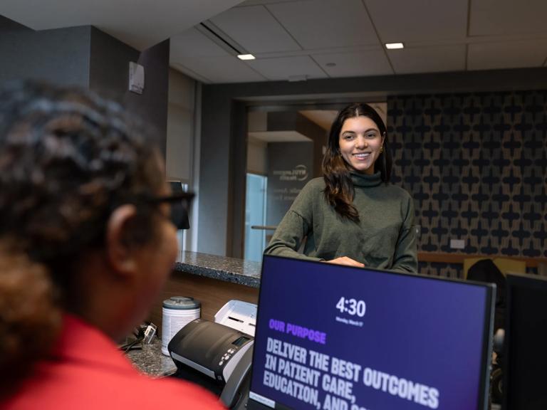 A female patient, left, stands at a check-in desk at a doctor's office and speaks to a receptionist, left, sitting in front of a computer monitor.