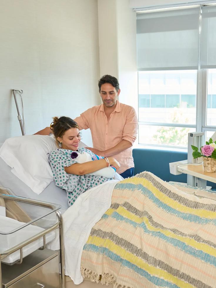 A female patient, left, lies in a hospital bed holding a baby while a man, right, stands next to her.