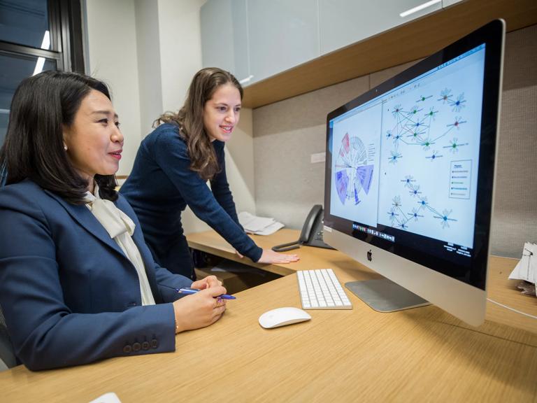 Two women are in an office looking at a computer monitor. One, left, is seated, while the other, right, stands.
