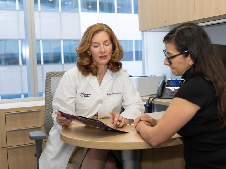 A female doctor sits behind a desk in an office. She looks at a folder with a female patient sitting across from her.