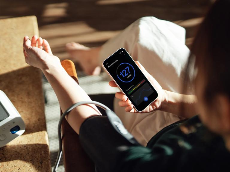 A person wearing a blood pressure cuff on their arm views a blood pressure reading on a smartphone health app while sitting at home.