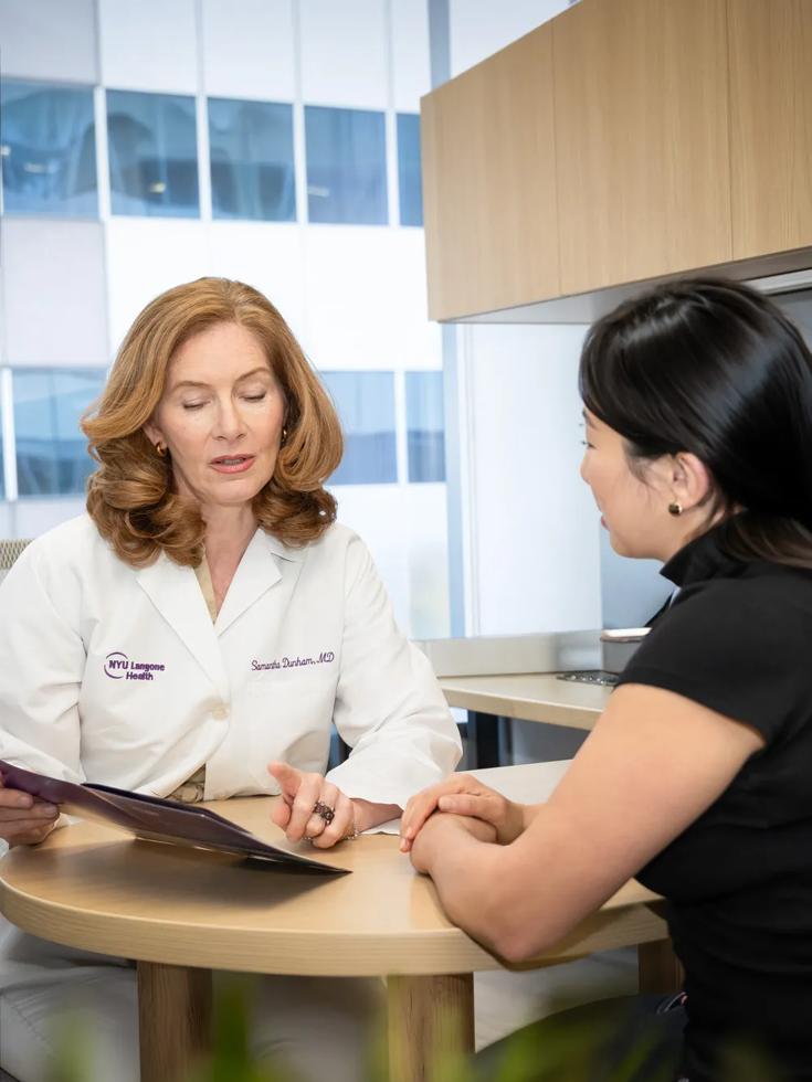 A female doctor sits behind a desk in an office. She looks at a folder with a female patient sitting across from her.