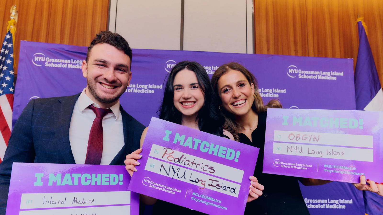 Three soon-to-graduate medical students from the Grossman Long Island School of Medicine smile and hold up pieces of paper that say "I Matched" in bold purple letters.