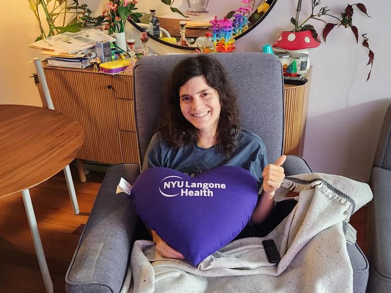 Shana Hale sits in a chair and holds a large purple pillow that&rsquo;s heart-shaped and reads &ldquo;NYU Langone Health.&rdquo;
