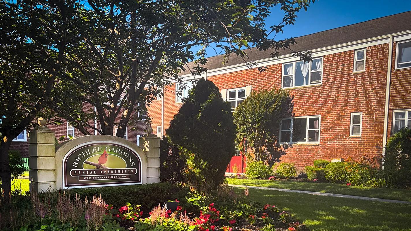 A two-story brick building that provides housing for NYU Grossman Long Island School of Medicine. In front of the building a cement sign, surrounded by flowers and shrubs, has the words "Richlee Gardens Rental Apartments" and a life-like image of a red cardinal on a green background.