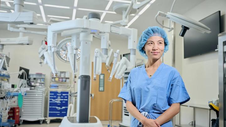 Kathy Huang, MD, stands in scrubs in an operating room with medical equipment in the background.