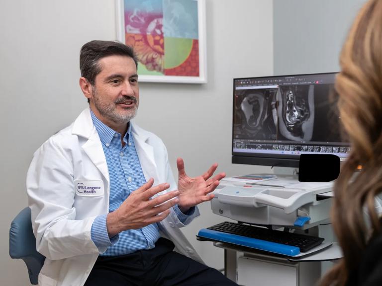 A doctor in an NYU Langone Health lab coat sits in an exam room, gesturing with his hands while explaining MRI scan images displayed on a computer monitor to a patient seated across from him.









