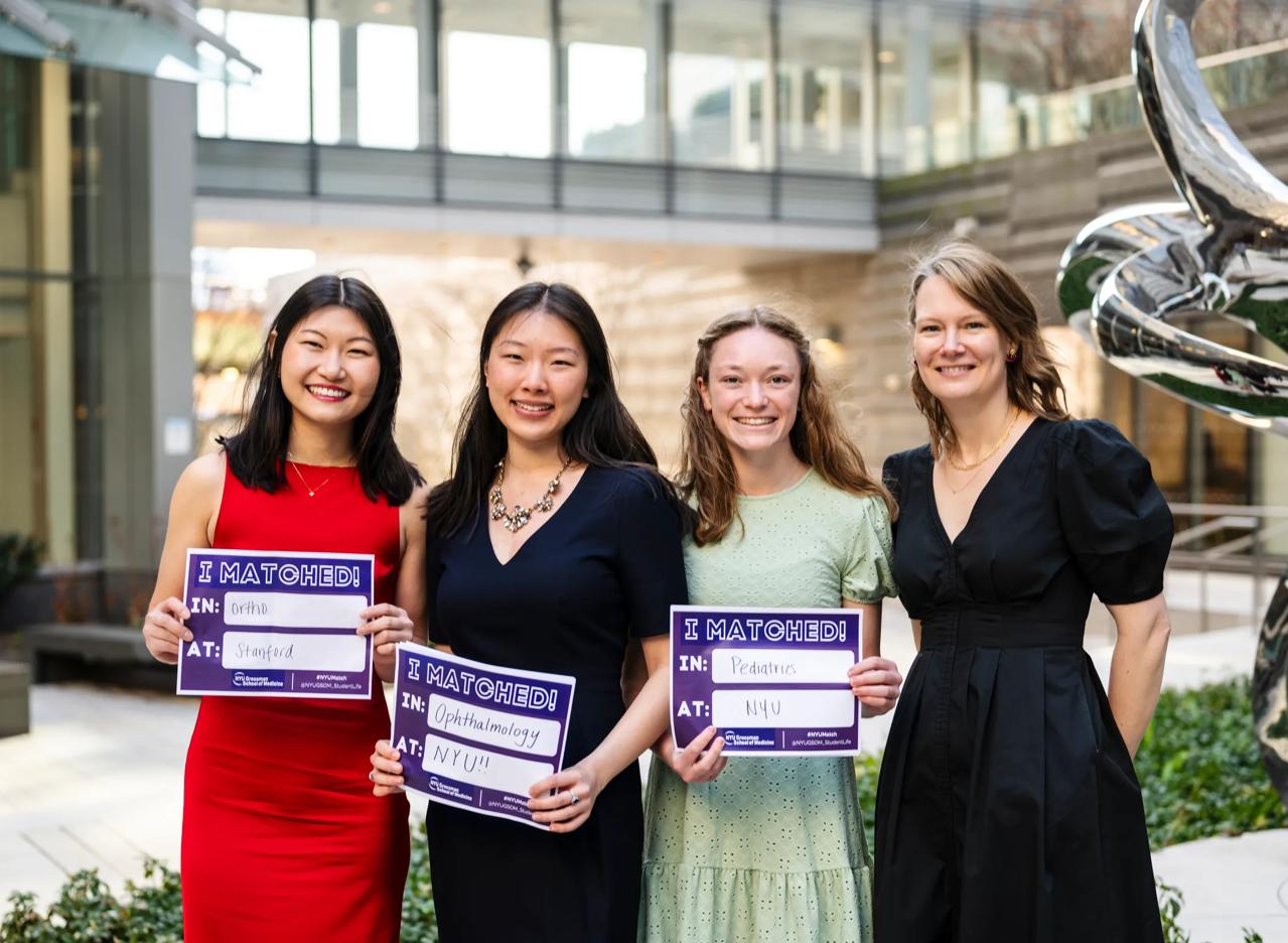 Melissa Song, Lily Ge, and Brooke Starn stand with the associate dean of student affairs, Dr. Victoria Dinsell during Match Day at the Grossman School of Medicine. Each is smiling, holding a sign listing their residency assignments. 