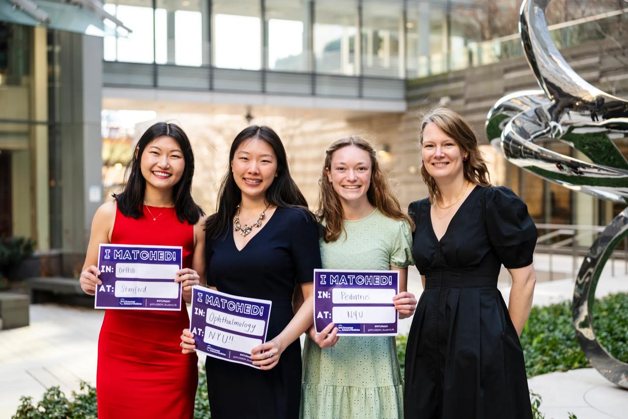 Melissa Song, Lily Ge, and Brooke Starn stand with the associate dean of student affairs, Dr. Victoria Dinsell during Match Day at the Grossman School of Medicine. Each is smiling, holding a sign listing their residency assignments. 