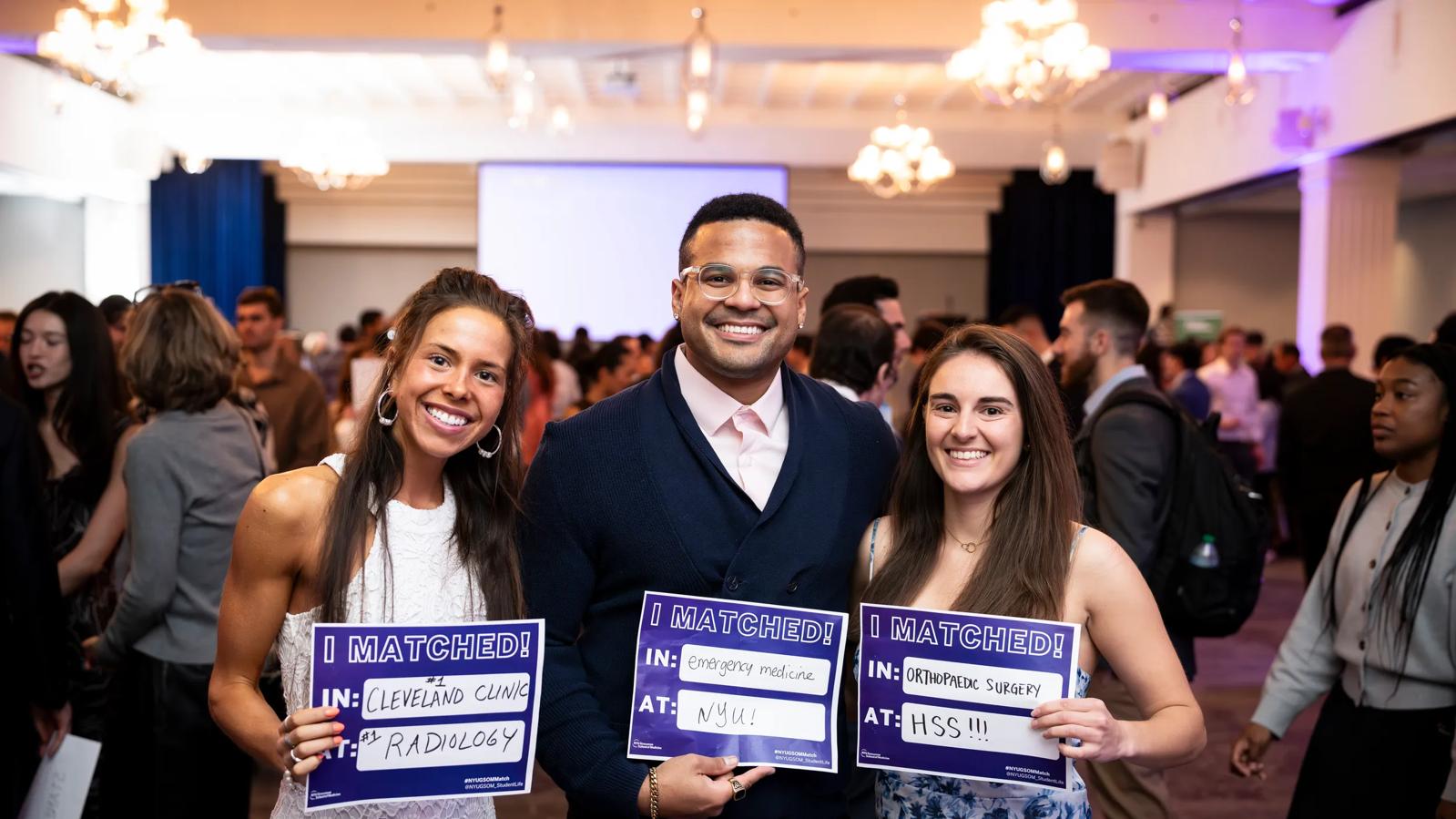Three soon-to-graduate medical students from the Grossman School of Medicine smile and hold up pieces of paper that say "I Matched" in bold purple letters.
