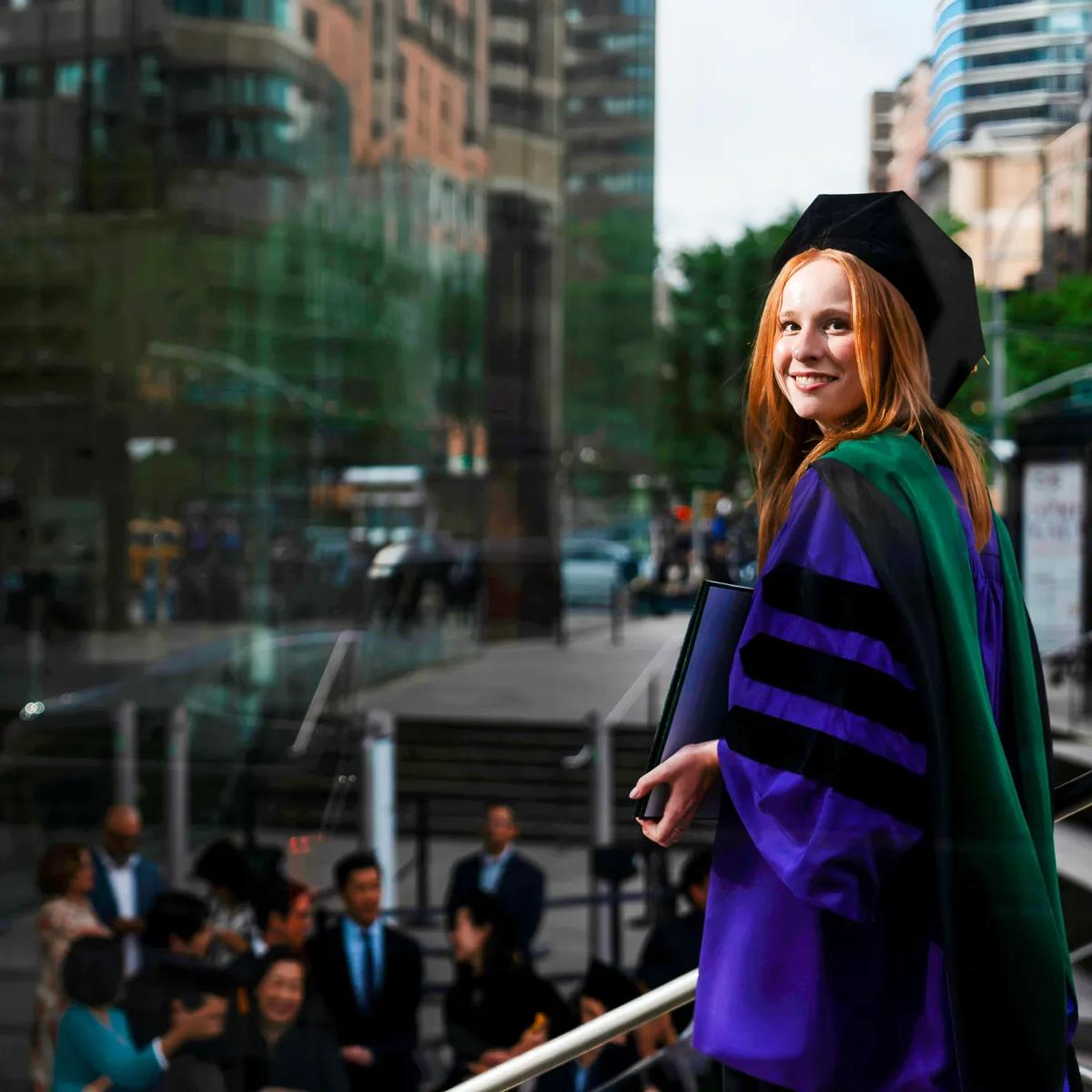 A female NYU Grossman School of Medicine graduate stands on a New York City sidewalk outside the school on a sunny day, smiling as she looks back at the camera with her purple commencement robe and doctoral hood.