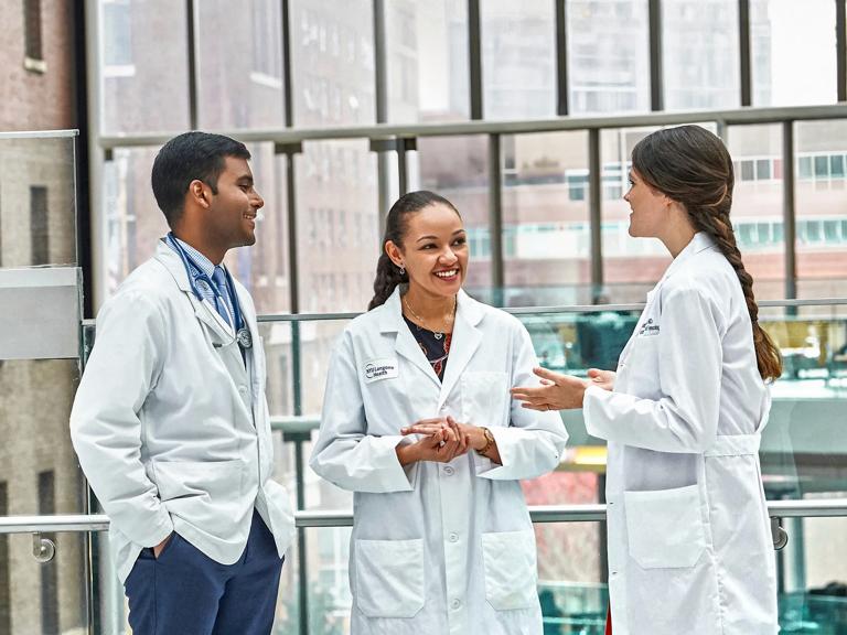 Three Grossman School of Medicine students wearing white coats smile as they talk in a hospital foyer.