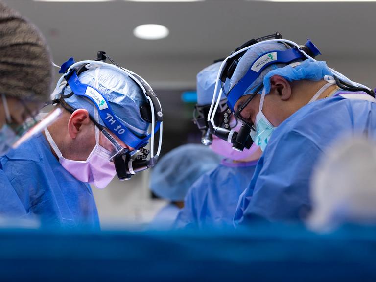Three doctors wearing protective gear stand in an operating room performing a transplant procedure.