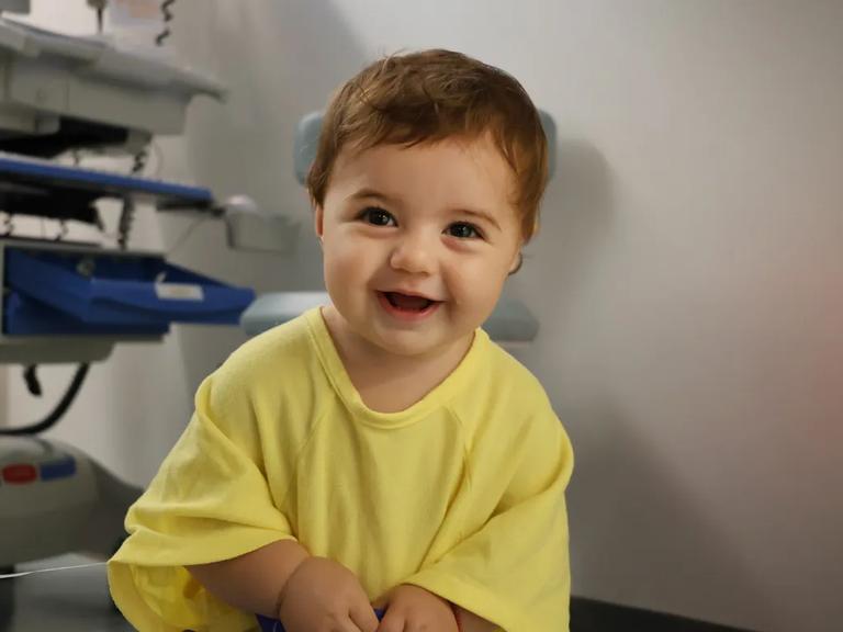 An 18-month-old girl with a cochlear implant smiles warmly while wearing a bright yellow T-shirt.