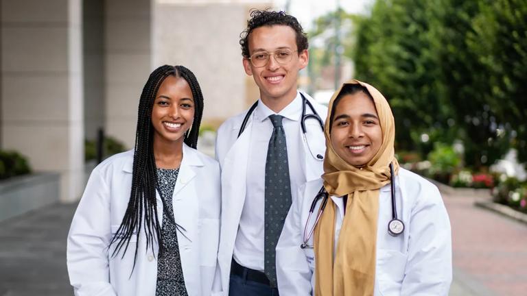 Three NYU Grossman Long Island School of Medicine students wearing white coats smile shoulder-to-shoulder standing outside the school.