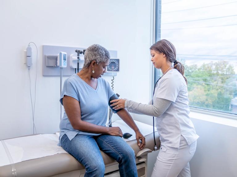 A female nurse, right, puts a medical cuff on the arm of a female patient, left, seated, to measure her blood pressure.