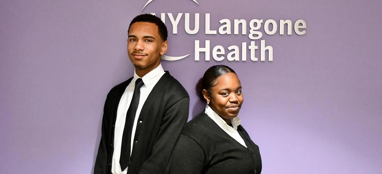 High school students Yadiel Antonio Santana and Esienee Morales stand in front of a purple wall with a  white NYU Langone Health logo.