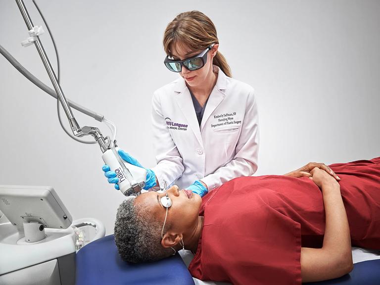 A female doctor, standing, performs a procedure on the forehead of a female patient, lying down.