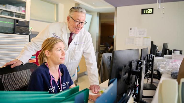 A male professor at NYU Grossman School of Medicine stands smiling over a desk whie his hand on a chair, while a female siting at a desk and wearing dark blue hospital scrubs uses a program on a computer.