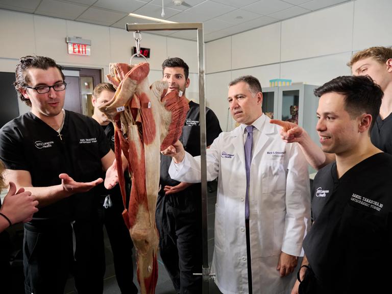 A male NYU Grossman Long Island School of Medicine professor wearing a white coat explains musculature to students studying orthopedic surgery wearing black hospital scrubs.