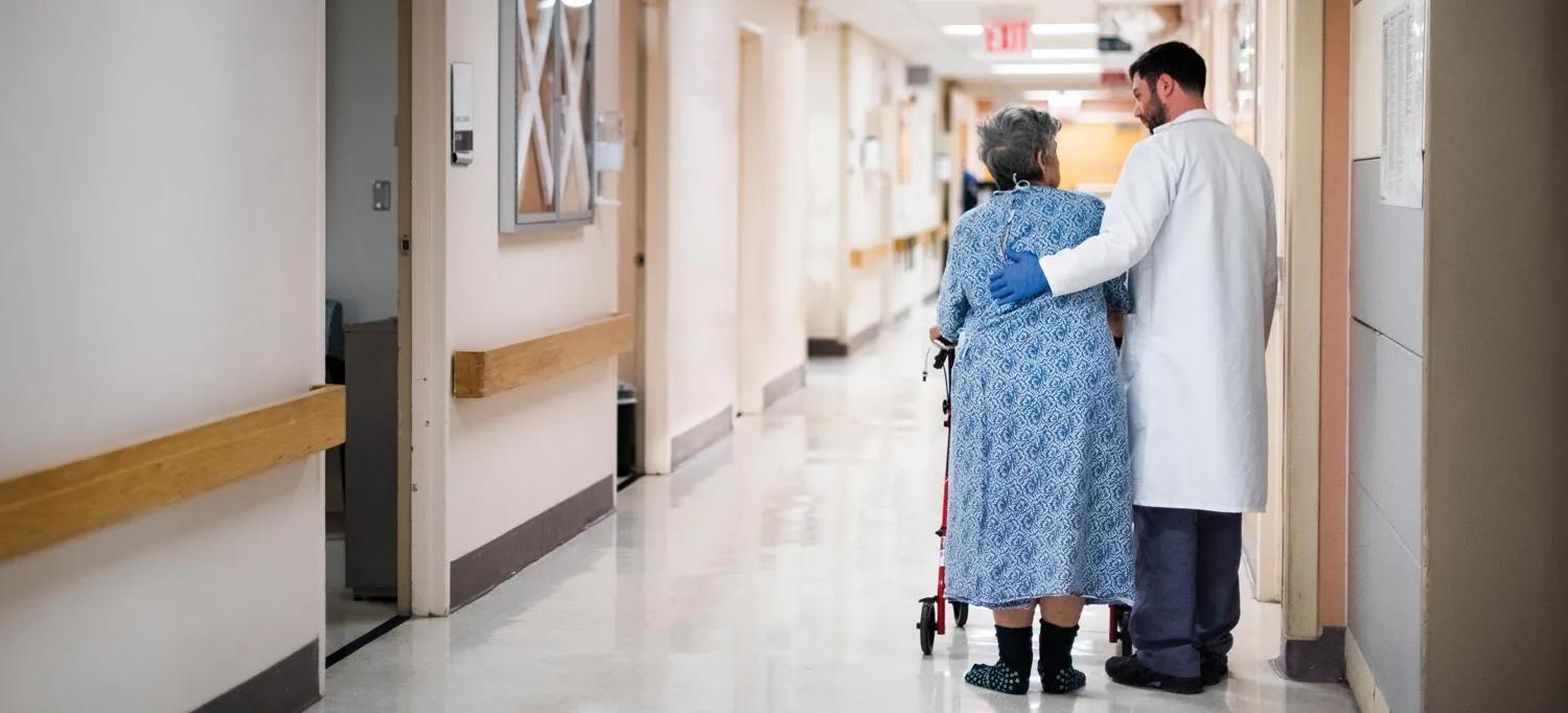 Doctor walking down a hospital hallway with an older patient using a walker, gently supporting them with an arm around their back