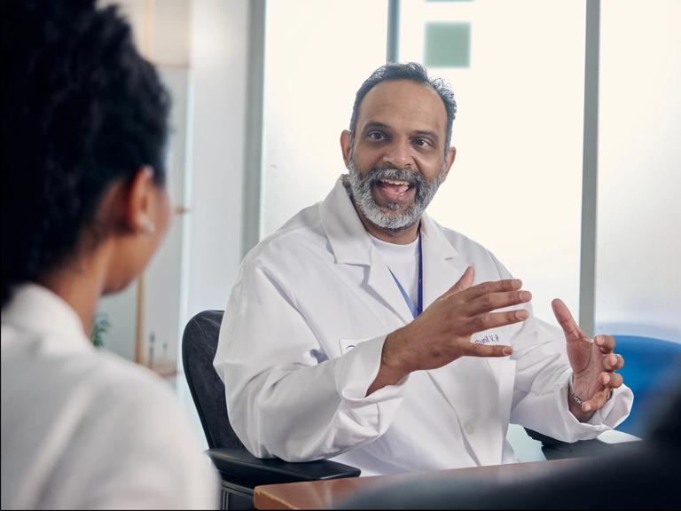 Sunil Rao, MD, gestures with his hands while sitting and speaking with a patient in a clinical office.