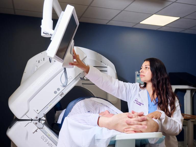 A female radiologist, left, stands while performing an imaging exam on a patient who is lying down with their hands above their head.