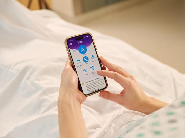 A person in bed holds a smartphone with an NYU Langone app open.