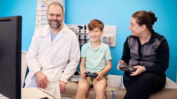 A male doctor, left, sits with a pediatric patient, center, and a woman, right, on an exam table looking at a monitor. The patient and woman hold video game controllers.