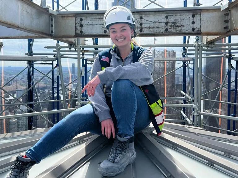 Wearing a hardhat and smiling, Oberle sits on a section of a building under construction.