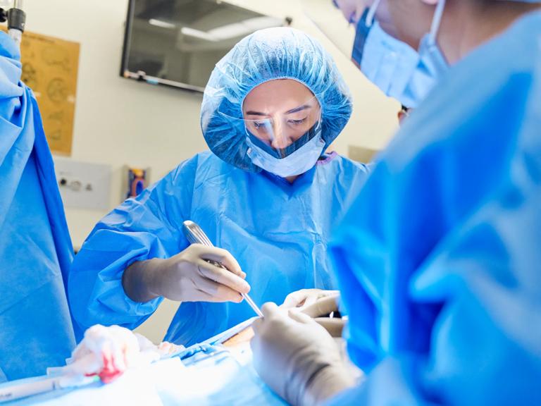 A female NYU Langone doctor wearing blue hospital scrubs, a mask, and a face shield, performs surgery on a patient.