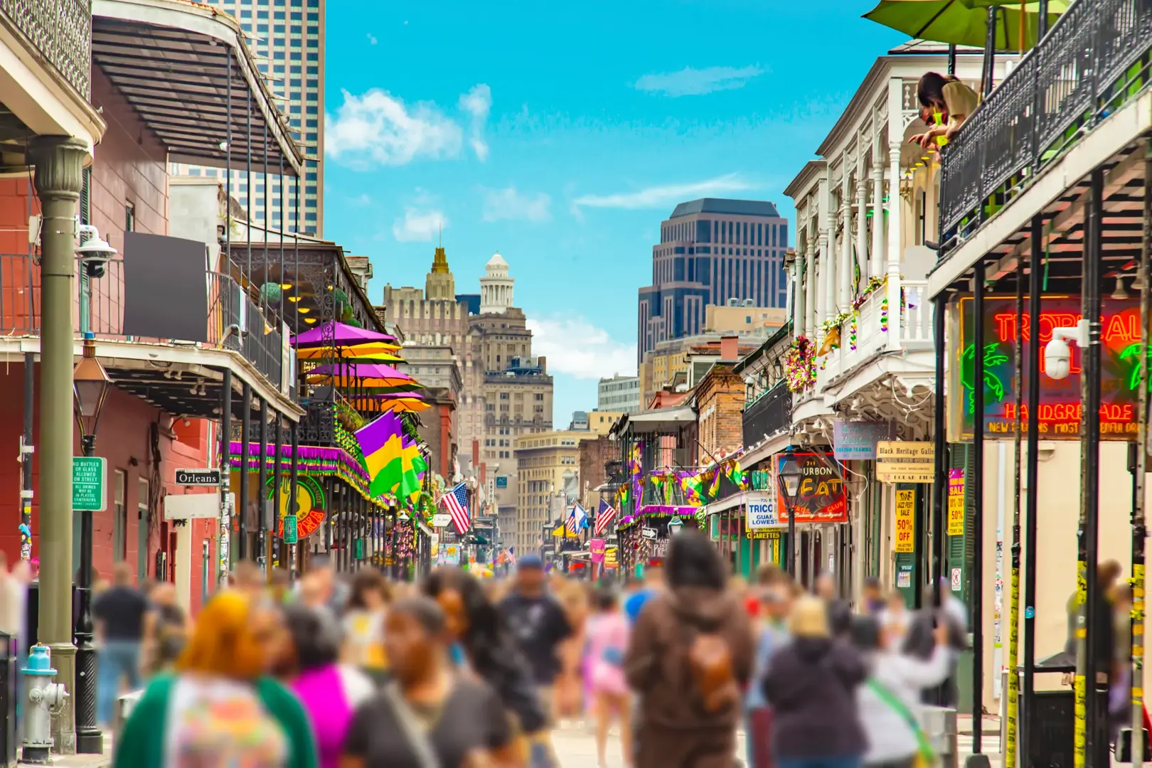 Crowds of people bustle along Bourbon Street in New Orleans, Louisiana, filling the vibrant thoroughfare with energy and excitement. The historic buildings lining the street are adorned in festive Mardi Gras colors—purple, green, and gold—creating a lively, celebratory atmosphere. In the background, the city’s skyline rises beneath a partly cloudy blue sky, adding a dynamic urban backdrop to the spirited scene.