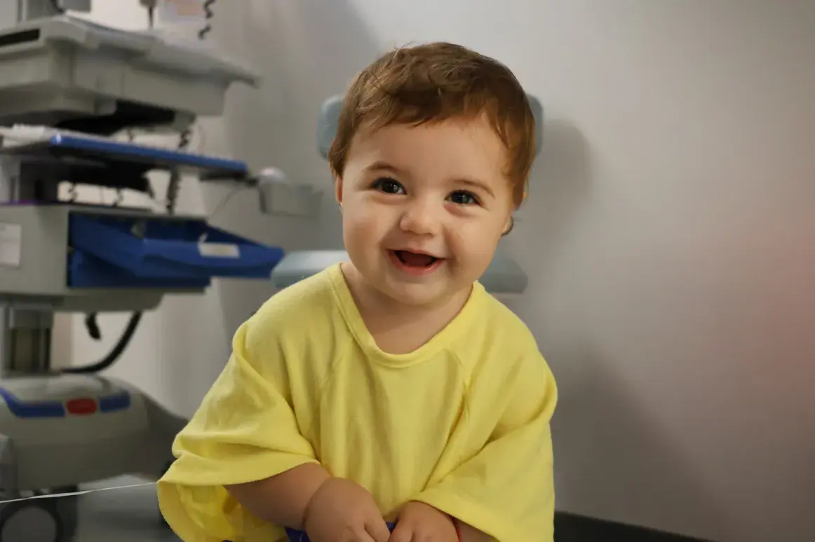 An 18-month-old girl with a cochlear implant smiles warmly while wearing a bright yellow T-shirt.