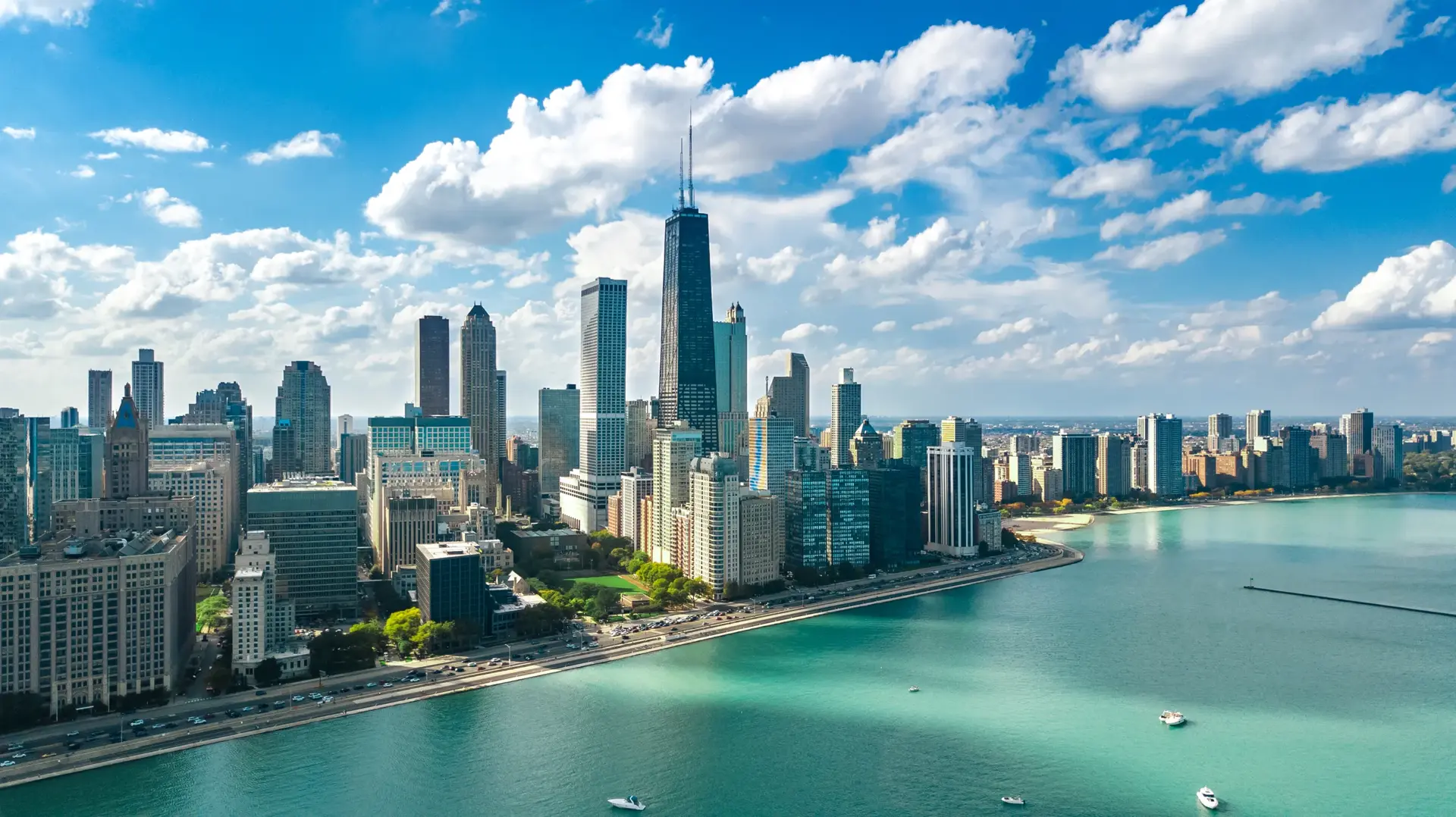 Aerial view of Chicago&rsquo;s skyline along Lake Michigan on a bright day, with the John Hancock Center rising above waterfront high-rises under a blue sky filled with scattered white clouds.









