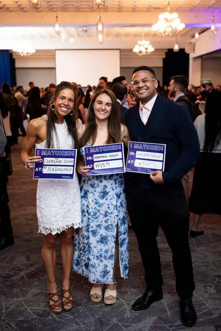 Two female NYU Grossman School of Medicine students in dresses next to a male classmate a suit, smiling while holding purple signs up that say in large letters "I Matched!"