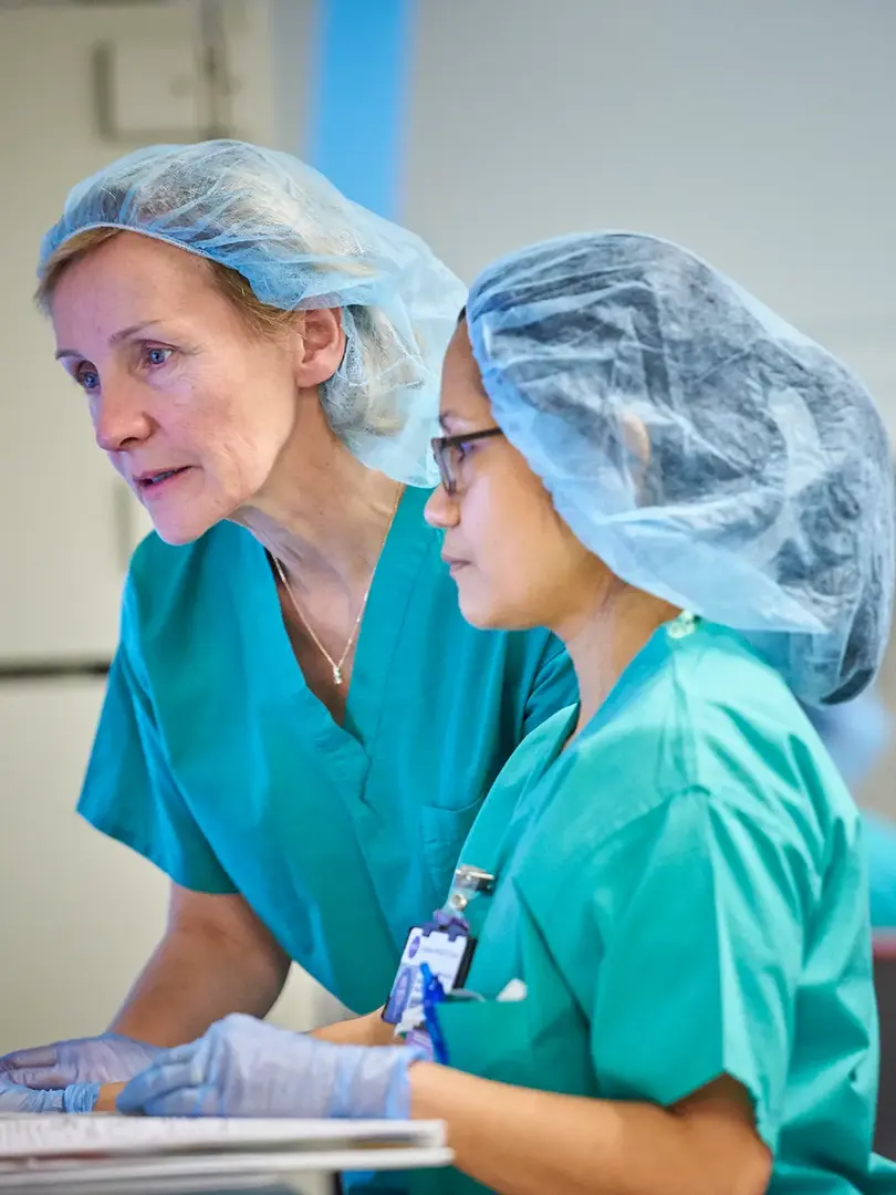 Two female nurses, standing, wear scrubs hair nets while looking at a monitor in a hallway.