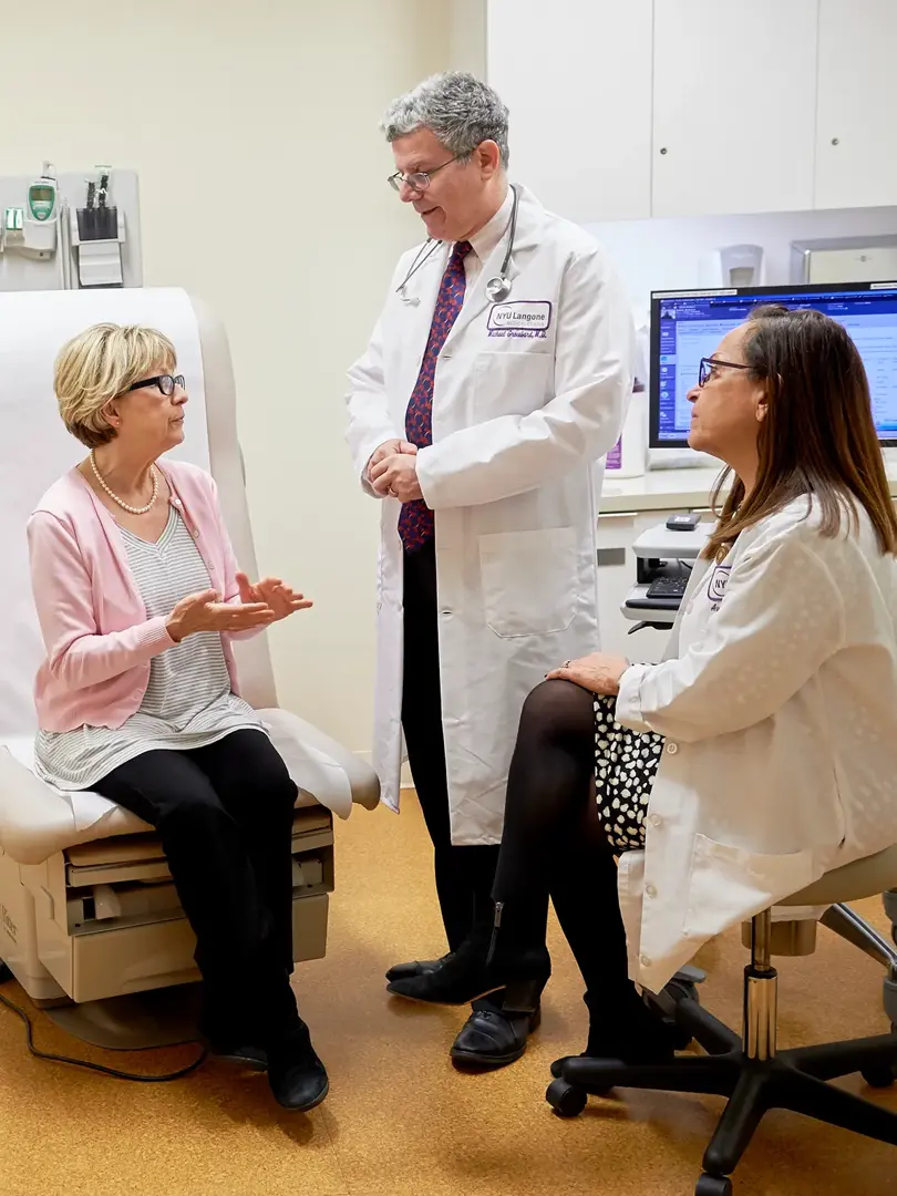 A female patient, left, sits in an exam room while speaking to a male doctor, center, standing, and a female doctor, right, sitting.