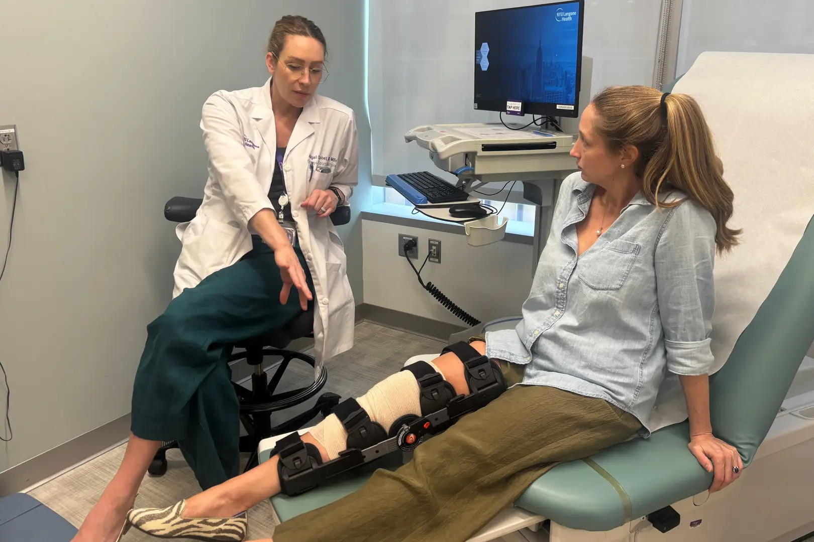 In a medical exam room, a healthcare professional in a white coat sits on a stool and gestures while talking with a patient who is seated on an exam table. The patient has a large black knee brace and bandage on one leg, which is extended onto a support.